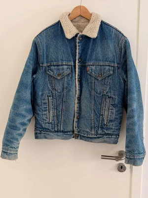 Levi's vintage blå jeansjacka med foder - Storlek 44  - Klassisk blå jeansjacka från Levi's med vit teddyfoder på insidan och kragen. Jackan har två bröstfickor med lock och knappar, samt två sidofickor. Rak passform och snygga detaljer som metallknappar och kontrastsömmar. Perfekt för dig som gillar tidlös streetstil.