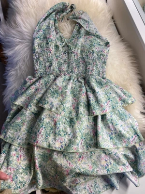 Blommig volangklänning med halterneck - Söt kortklänning med blommigt mönster i ljusa pastellfärger. Klänningen har halterneck, smockad överdel och flera lager volanger i kjolen. Perfekt för sommarens alla tillfällen!