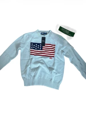 Ljusblå stickad tröja Polo Ralph Lauren - Ljusblå stickad tröja från Polo Ralph Lauren med amerikansk flagga broderad på bröstet och RL-detalj i rött. Tröjan har rund halsringning och ribbade muddar vid ärmslut och nederkant. Perfekt för dig som gillar klassisk preppy stil.