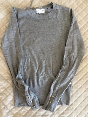 Grå stickad tröja med knappar - Snygg grå stickad tröja från lindex med rund halsringning och ribbade muddar. Ärmsluten har dekorativa guldknappar som ger en extra detalj. Perfekt till jeans eller kjol för en clean look. 