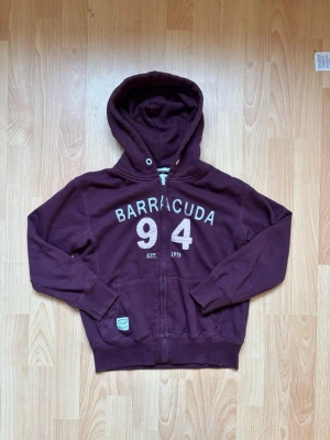 Vinröd hoodie från Barracuda - Snygg vinröd hoodie med dragkedja från Barracuda. Tryck med texten 'BARRACUDA 94 EST. 1979' på bröstet. Hoodien har huva med dragsko, ribbade muddar och två fickor framtill. Perfekt för en avslappnad och sportig stil.