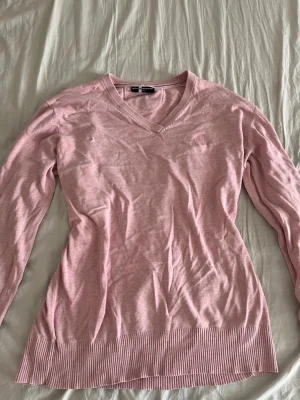 Ljusrosa stickad tröja Gant - Ljusrosa stickad tröja från sail racing med v-ringning och diskret broderad logga på bröstet. Tröjan har långa ärmar och ribbstickade muddar vid ärmslut och nederkant. Perfekt för lager-på-lager och en avslappnad stil.