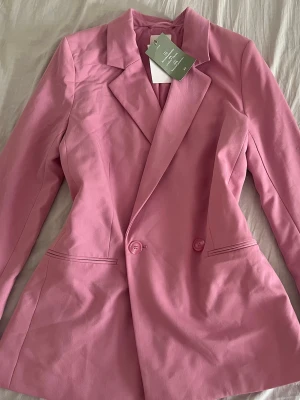 Rosa kavaj med breda slag - Snygg rosa kavaj med breda slag och två knappar framtill. Kavajen har figursydd passform och två fickor framtill. Perfekt för dig som vill sticka ut med färg. Materialet känns mjukt och har en lätt stretch.