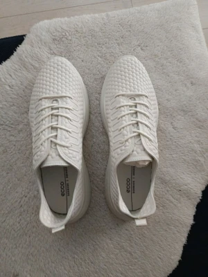 Vita sneakers från ECCO med vävt mönster - Snygga vita sneakers från ECCO med ett unikt vävt mönster över hela skon. Skorna har vita snören och en låg profil. Tillverkade i ett mjukt material som ger en modern och clean look. Perfekta för dig som gillar stilrena och bekväma sneakers.