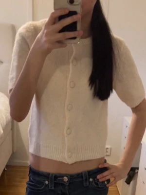 Beige kortärmad kofta med knappar - Söt beige stickad kofta med korta ärmar och rund hals. Koftan har ribbade kanter och knäppning framtill med matchande knappar. Perfekt att styla över ett linne eller klänning för en trendig look.