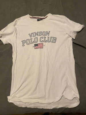 Vit t-shirt Vinson Polo Club - Vit t-shirt från Vinson Polo Club med tryck framtill och amerikansk flagga. Klassisk rund halsringning och korta ärmar. Perfekt till jeans eller shorts för en avslappnad look.