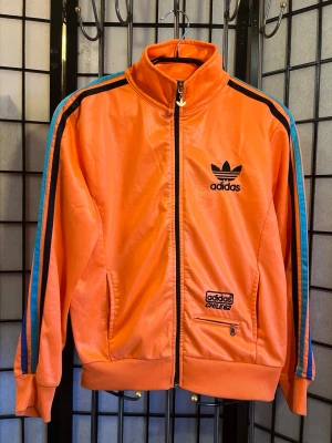 🔥 Adidas Chile 62 Track Jacket – Orange / Trefoil / Rare 🔥 - 🔥 Adidas Chile 62 Track Jacket – färgbomb 💥 Glossy finish + stor trefoil bak = riktig statement piece. Retro/Y2K vibe som sticker direkt.  • Chile 62 • Broderad trefoil fram • Stor multi-color trefoil bak • Zip pockets + trefoil zipper  Stl L (liten – sitter som S/M, fitted modell) Mått: • Pit to pit (armhåla–armhåla): 48–49 cm • Längd (axel till nederkant): 56–57 cm • Axel till axel: 41–42 cm  Skick: Nära nyskick • 2 små prickar • I övrigt typ oanvänd  699 kr – bud ok