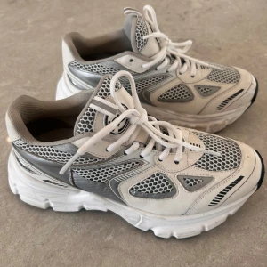 Vit och grå sneaker från Axel Arigato - Snygga sneakers från Axel Arigato i vitt och grått med meshpaneler och detaljer i syntetmaterial. Skorna har tjock sula, klassisk snörning och logotyp på hälen. Perfekt för dig som gillar en sportig och modern look.