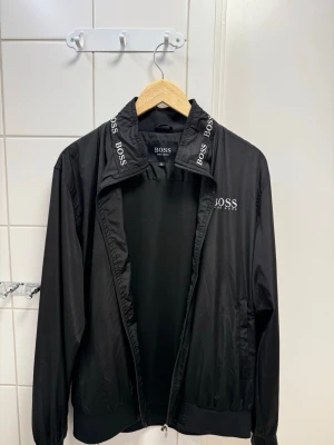Svart vindjacka från BOSS - Snygg svart vindjacka från BOSS med dragkedja och hög krage. Jackan har vita BOSS-loggor på kragen och bröstet, samt två sidofickor. Tillverkad i lätt och tunt material som passar perfekt till blåsiga dagar.