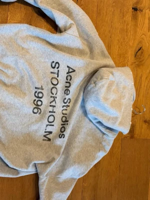 Grå hoodie Acne Studios Stockholm - Säljer nu denna eftertraktade gråa hoodien från Acne Studios med trycket 'Acne Studios STOCKHOLM 1996' på ryggen. Acnes kläder är lite större i storlek så om du är en M passar denna jättebra, perfekt för en avslappnad streetwear-look. Enkel design utan fickor framtill.