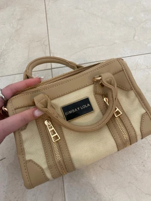 Beige handväska från Bimba Y Lola - Snygg beige handväska från Bimba Y Lola i canvas och läderdetaljer. Väskan har guldfärgade dragkedjor, dubbla handtag och en avtagbar axelrem. Märkesplakett framtill och flera fack inuti för smart förvaring.