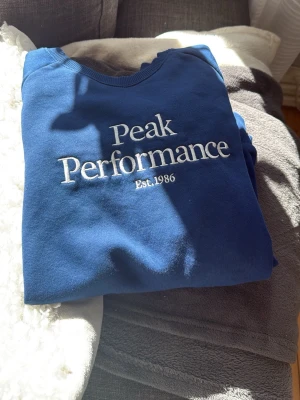 Blå sweatshirt från Peak Performance - Snygg blå sweatshirt från Peak Performance med vit broderad logga och texten 'Est. 1986' på bröstet. Tröjan har rund halsringning, ribbade muddar vid ärmslut och nederkant. Perfekt för en avslappnad och sportig stil.
