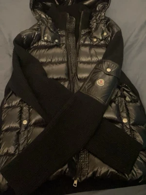 Svart dunväst från Moncler - Snygg svart dunväst från Moncler med glansigt quiltat material och stickade svarta ärmar. Västen har huva, dragkedja framtill och silverfärgade knappar. Moncler-logga på ärmen och praktiska fickor. Perfekt för att hålla sig varm och stilren. ÄKTA!