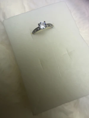 Silverfärgad ring med klar sten - En elegant silverfärgad ring med en stor, klar rund sten i mitten och mindre gnistrande stenar längs bandet. Ringen har en klassisk och stilren design som passar dig som gillar att sticka ut med snygga accessoarer. Perfekt för att ge din look lite extra glans.