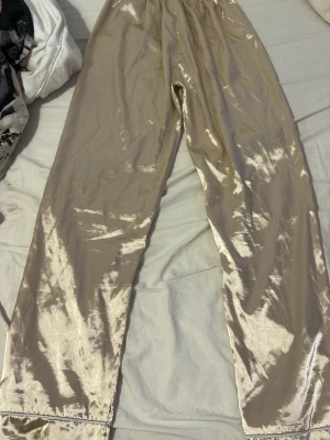 Champagnefärgad satinpyjamas - Snygg tvådelad pyjamas i glansigt champagnefärgat satintyg. Setet består av långärmad skjorta med krage och knappar samt matchande byxor med rak passform. Perfekt för mysiga kvällar hemma.