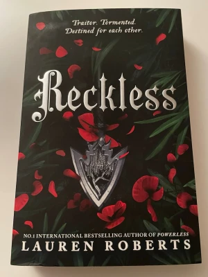 Reckless av Lauren Roberts - En spännande skönlitterär bok med svart omslag dekorerat med röda blomblad och ett silverfärgat vapenmotiv. Titeln 'Reckless' är tryckt i stora vita bokstäver och boken är skriven av Lauren Roberts, känd för Powerless. Perfekt för dig som gillar fantasy och romantik. Har bara ståt på bokhyllan, aldrig läst 
