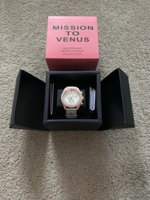 Omega x Swatch MoonSwatch Venus - Säljer en Omega x Swatch MoonSwatch Mission to Venus med ljusrosa boett och vit urtavla. Klockan har tre subdials, tachymeter och vit rem med Omega-logga. Biokeramisk material och modern design, perfekt för dig som gillar unika accessoarer.