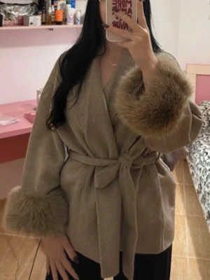 Havregrynsfärgad kappa med stora pälspuffar - Stilren kappa med knytskärp i midjan och breda ärmslut dekorerade med fluffig tjock päls. Kappan har en avslappnad passform och är perfekt för dig som vill ha en trendig och mysig look. Materialet ser ut att vara mjukt och värmande ullblandning.