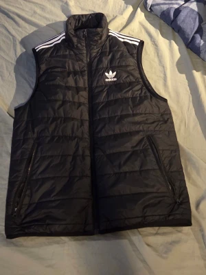 Svart dunväst från Adidas Originals - Svart dunväst från Adidas Originals med vita ränder på axlarna och klassisk logga på bröstet. Västen har hög krage, två dragkedjeförsedda fickor och quiltad design. Perfekt för lager-på-lager och streetwear-stil.