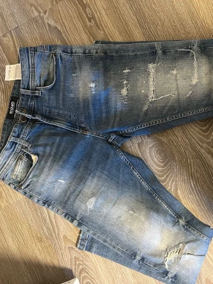 Blå slitna jeans från Iconic - Säljer ett par blå jeans från Iconic med snygga slitningar och distressed detaljer. Jeansen har fem fickor, normal midja och raka ben. Perfekta för en avslappnad och trendig look. Materialet är klassisk denim i bomull.