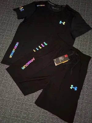 Svart träningsset från Under Armour - Svart träningsset från Under Armour med färgglad logga och textdetaljer i regnbågsfärger. Shortsen har snörning i midjan och en sportig look, perfekta för träning eller chill. Matchar med t-shirt i samma stil. Storlek S, helt ny 