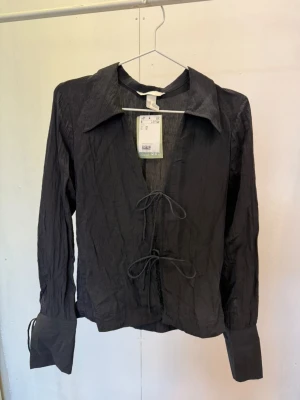 Svart crinkle-blus med knytning - Trendig svart blus i crinkle-material med två knytband framtill och bred krage. Långärmad modell med vida manschetter och avslappnad passform. Perfekt för dig som gillar en edgy och modern stil.