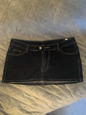 Svart jeanskjol med kontrastsömmar - Snygg mörk marinblå kort jeanskjol med tydliga vita kontrastsömmar och klassiska  fickor. Kjolen har bälteshällor och knappstängning framtill. Köpt på en marknad i Spanien och är HELT NY men för kort för mig. Passar S och M!