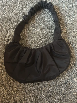 Svart axelremsväska med rynkad rem - Trendig svart handväska i nylon med rynkad axelrem och mjuk, rundad form. Väskan har en enkel men cool design som passar perfekt till streetwear och casual outfits. Perfekt storlek för det viktigaste och lätt att matcha med olika stilar.