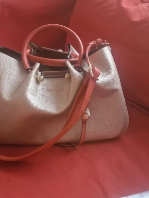Beige handväska - Snygg beige handväska med röda handtag och axelrem. Väskan har en strukturerad yta i konstläder och guldfärgade metalldetaljer. Perfekt för dig som vill ha en stilren och trendig accessoar.