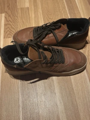 Bruna sneakers från Rieker - Snygga bruna sneakers från Rieker med svarta detaljer och svarta skosnören. Skorna har en ovandel i läder och textil, samt en bekväm innersula med Evolution Soft-teknologi. Yttersulan är ljus och ger en schysst kontrast till resten av skon.