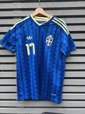 Sverige fotbollströja Adidas blå - Blå fotbollströja från Adidas med Sveriges landslagsemblem och nummer 17 i gult. Tröjan har gula ränder på axlarna, V-ringning och mönstrad design. Namnet Gyökeres står på ryggen. Perfekt för dig som vill stötta Sverige på match!