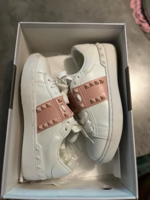 Valentino Rockstud Untitled sneakers - Snygga vita sneakers från Valentino med rosa band och ikoniska pyramidnitar över vristen. Skorna har en klassisk låg modell, vita snören och detaljer i läder. Perfekta för dig som vill sticka ut med en lyxig och trendig look. Använd endast en gång