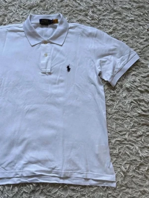 Vit pikétröja från Polo Ralph Lauren - Klassisk vit pikétröja från Polo Ralph Lauren med korta ärmar, krage och knappar framtill. Tröjan har den ikoniska broderade loggan på bröstet och är tillverkad i mjuk bomull. Perfekt för en clean och stilren look.