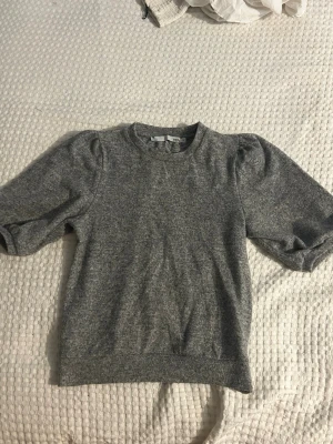 Grå tröja med puffärm från 157 - Mysig grå tröja från 157 med rund halsringning och trendiga puffärmar. Tröjan har en enkel och stilren design, perfekt att matcha med jeans eller kjol. Mjuk och bekväm, passar dig som gillar en avslappnad men ändå snygg look.
