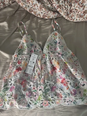Blommig broderad linnetopp Gina Tricot - Söt linnetopp från Gina Tricot med blommigt mönster i flera färger. Toppen har tunna axelband, broderade detaljer och genombruten spets. Perfekt för sommarens varma dagar och ger en romantisk vibe.