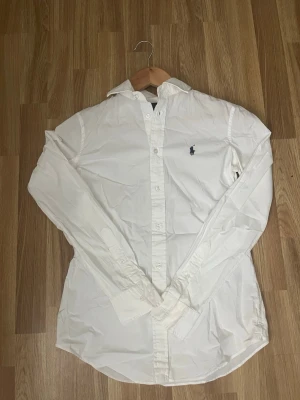 Vit skjorta från Ralph Lauren - Klassisk vit skjorta från Ralph Lauren med lång ärm och knappar framtill. Skjortan har den ikoniska broderade loggan på bröstet och är tillverkad i bomull. Perfekt till både jeans och kostymbyxor.