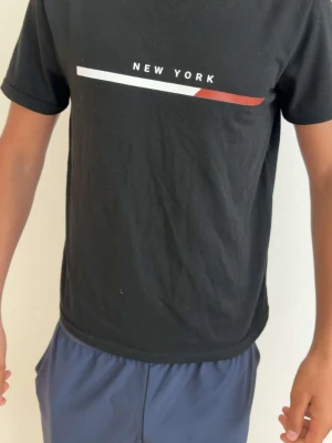 Svart t-shirt med New York-tryck - Svart t-shirt med korta ärmar och tryck framtill där det står 'NEW YORK' samt en vit och röd linje. Enkel och stilren design, perfekt till vardags. T-shirten har en normal passform och är troligtvis i bomull.