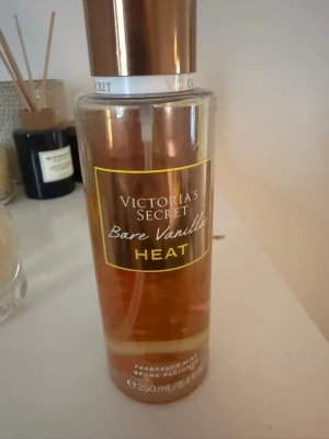 Victoria's Secret Bare Vanilla Heat - En doftmist från Victoria's Secret i varianten Bare Vanilla Heat. Flaskan är genomskinlig med guldiga och bruna detaljer och rymmer 250 ml. Doften har en varm och söt vaniljton, perfekt för dig som gillar mjuka och sensuella parfymer.