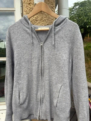 Kashmir Zip Hoodie - Mysig grå hoodie med dragkedja framtill och två fickor. Tillverkad i mjukt material och har en klassisk huva med dragsko. Perfekt för chill dagar eller när du vill ha en enkel och stilren look.