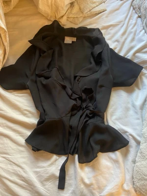 Svart omlottopp med volang - Säljer en svart omlottopp från asos med volangkant och knyt i midjan. Toppen har korta ärmar och en v-ringad urringning som ger en snygg och feminin look. Perfekt att styla med både jeans och kjol.