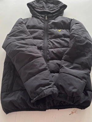 Svart pufferjacka från Lyle & Scott - Säljer en svart pufferjacka från Lyle & Scott med huva och dragkedja framtill. Jackan har det klassiska gula logomärket på bröstet och är quiltad för extra värme. Tillverkad i slitstarkt syntetmaterial och har elastiska muddar vid ärmslut och nederkant.