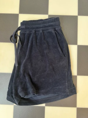 Mörkblå frottéshorts med snörning från Arket - Mysiga mörkblå shorts i mjuk frotté från Arket med hög midja och snörning i midjan. Shortsen har två sidofickor och en avslappnad passform, perfekta för varma dagar.