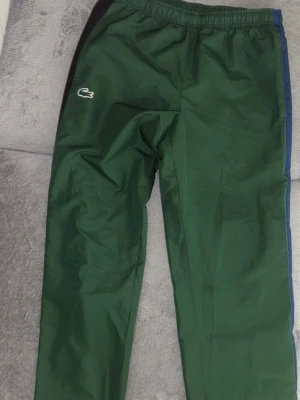 Gröna mjukisbyxor från Lacoste - Mörkgröna mjukisbyxor från Lacoste med elastisk midja och det klassiska krokodilmärket på vänster ben. Byxorna har en svart panel på sidan och blå detalj längs benet. Perfekta för en avslappnad och sportig stil.