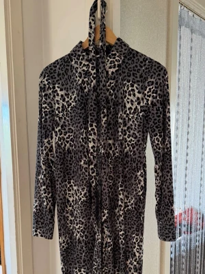 Leopardmönstrad klänning från JDY - Säljer en snygg leopardmönstrad klänning från JDY i svart och grått. Klänningen har lång ärm, knappar framtill och ett matchande knytband i halsen. Perfekt för dig som vill sticka ut lite extra med ett djurmönster.