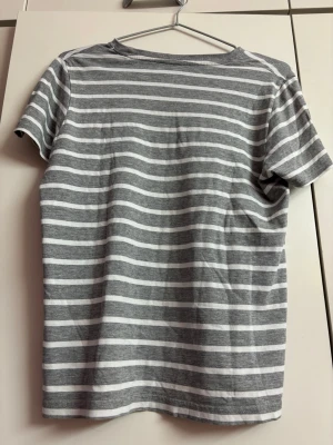 Gråvit randig t-shirt  - En stilren t-shirt med grå och vita horisontella ränder. Klassisk rund halsringning och korta ärmar. Perfekt för en avslappnad look och enkel att matcha med jeans eller shorts.