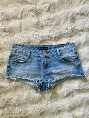 Ljusblå jeansshorts med fransig kant - Säljer ett par ljusblå jeansshorts från Bik Bok med fransig kant och slitna detaljer. Klassisk femficksmodell med knapp och dragkedja framtill. Perfekta för varma sommardagar och enkel att matcha med crop tops eller t-shirts.
