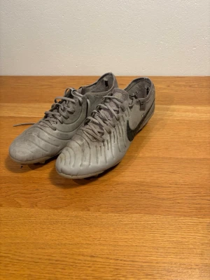 Gråa Nike tiempo fotbollsskor  - Säljer ett par gråa fotbollsskor från Nike med klassisk svart swoosh på sidan. Skorna har snörning, låg profil och dobbar för bra grepp på planen. Tillverkade i syntetmaterial med textilpartier för extra komfort. Perfekta för dig som vill ha stilrena och funktionella skor.