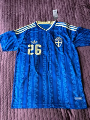 Blå Sverige fotbollströja Adidas - Säljer en blå Sverige fotbollströja från Adidas med gula detaljer och nummer 26. Tröjan har klassiska Adidas-ränder på axlarna, broderad landslagssköld och namn samt nummer på ryggen. Tillverkad i lätt och ventilerande material, perfekt för match eller träning.