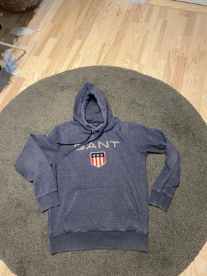 Blå hoodie från GANT med logga - Snygg blå hoodie från GANT med stor vit logga och sköld på bröstet. Klassisk känguruficka framtill och justerbar huva med dragsko. Mjuk bomullsblandning som är skön att ha på sig. Perfekt för en avslappnad och sportig stil.