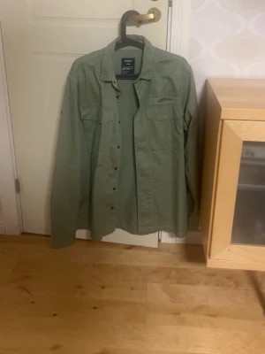 Grön skjorta från Superdry - Snygg grön skjorta /jacka från Superdry med klassisk krage och knappar framtill. Skjortan har två bröstfickor och är långärmad, perfekt för en avslappnad men stilren look.